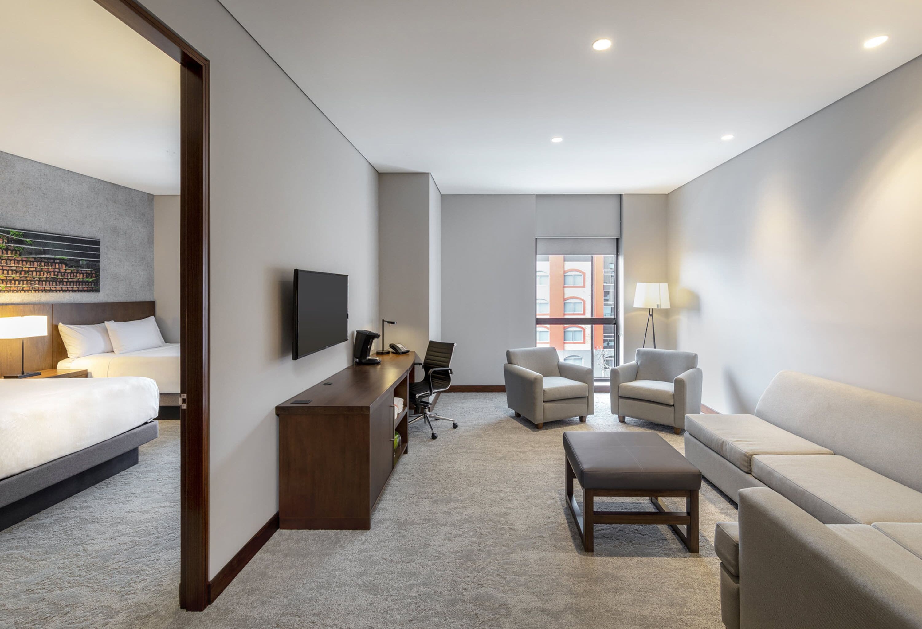 Una habitación de hotel moderna con una cama, un escritorio, un televisor, dos sillones, un sofá y una ventana con vista a la ciudad.