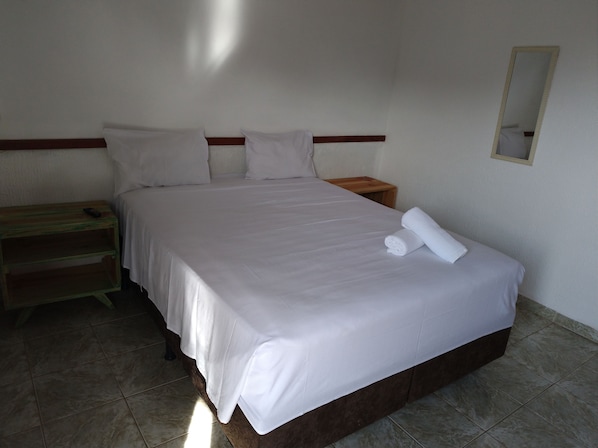 A neatly made bed with white linens, a wooden nightstand with a remote control, and a wall-mounted mirror.