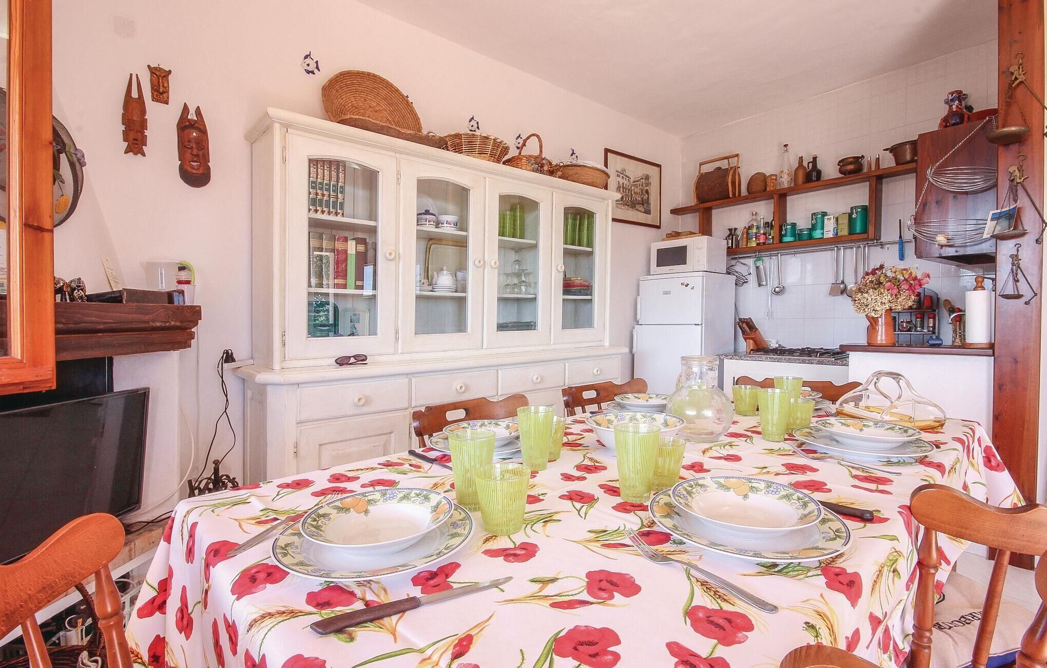 Un comedor con una mesa dispuesta para cuatro personas, rodeada de sillas. Un armario blanco con puertas de vidrio está contra la pared, y se observa una zona de cocina al fondo.