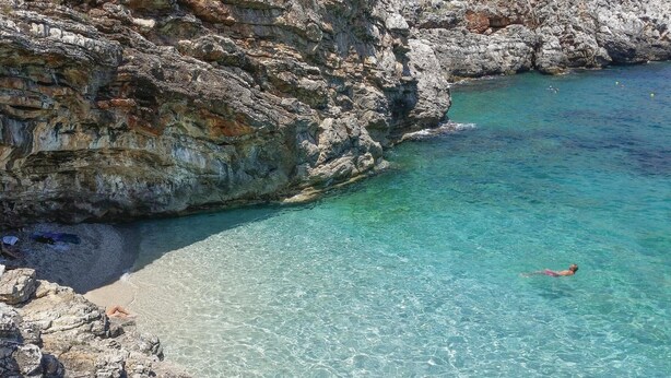 Costa rocosa con agua turquesa cristalina, un nadador y una playa pedregosa.