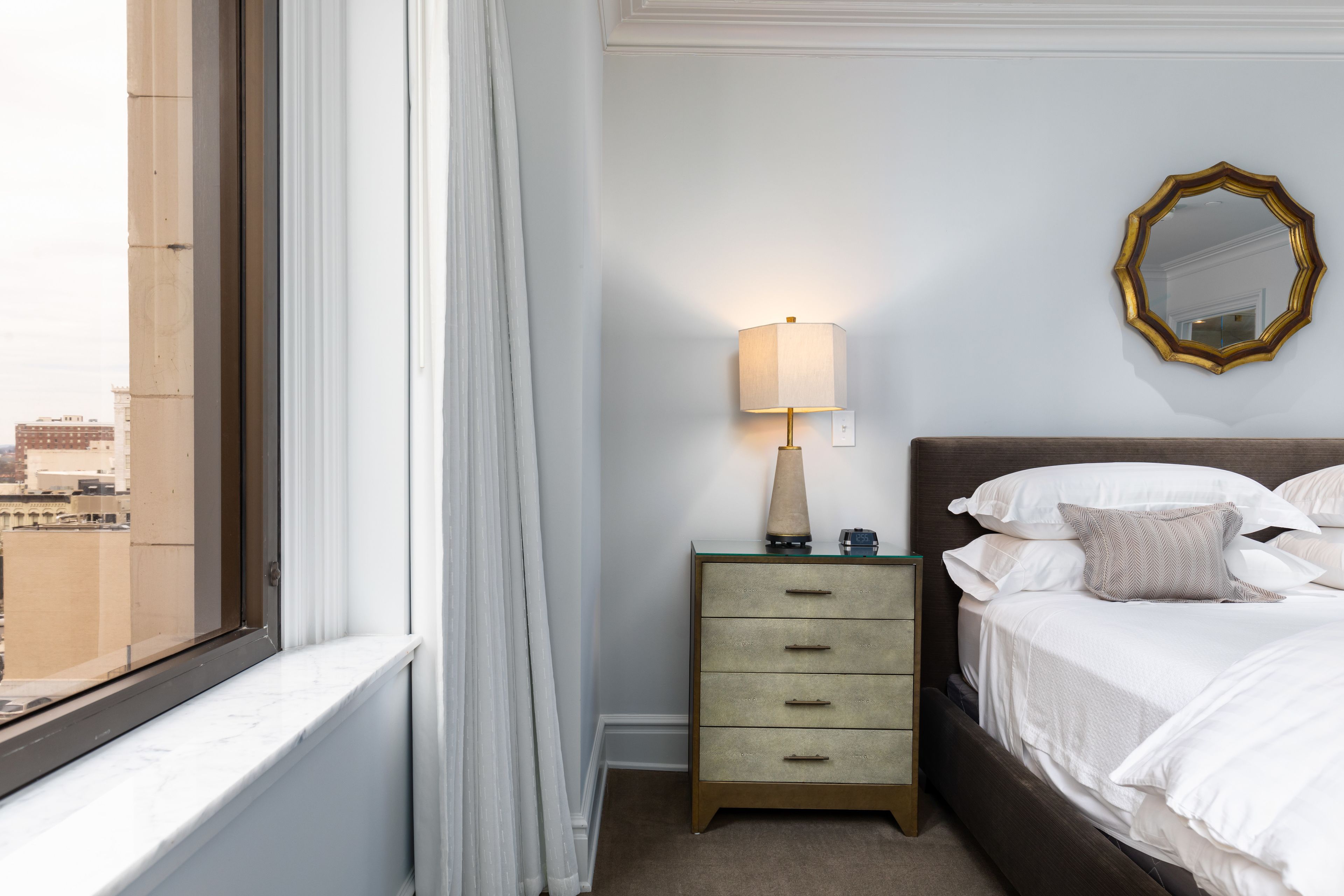 Een hotelkamer met een bed, nachtkastje met lamp, spiegel en uitzicht op de stad door een raam.