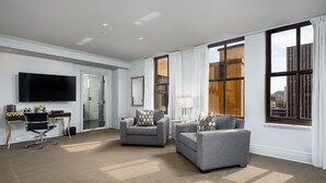 A modern hotel room with a large flat-screen TV, two grey sofas, a desk with a computer, and large windows with curtains. - John Hand Club Hotel (Birmingham)