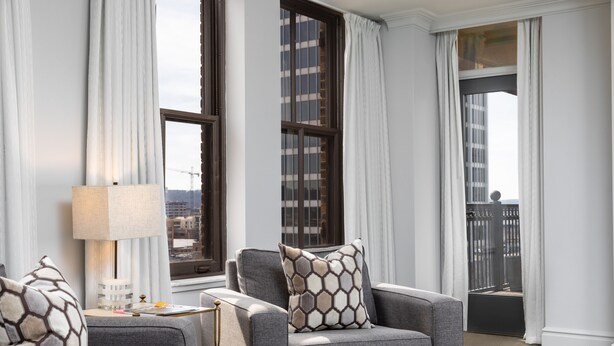 A modern living room with grey sofas, a side table, and a lamp. There are large windows with curtains, and a view of buildings outside. - John Hand Club Hotel (Birmingham)