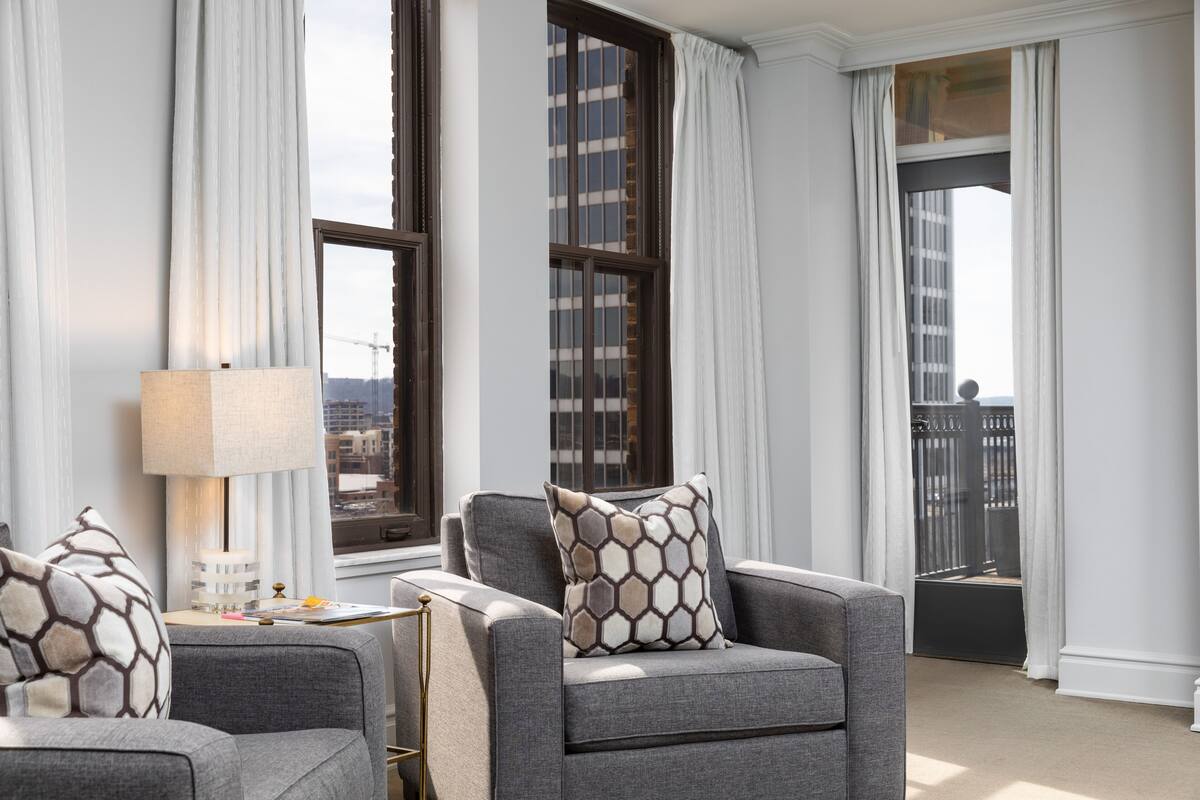 Una sala de estar moderna con sofás grises, una mesita auxiliar y una lámpara. Hay ventanas grandes con cortinas y vistas a edificios.