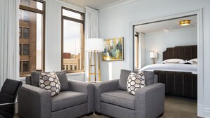 A hotel room with two grey armchairs, a bed, a painting, and a view of buildings through large windows. - John Hand Club Hotel (Birmingham)