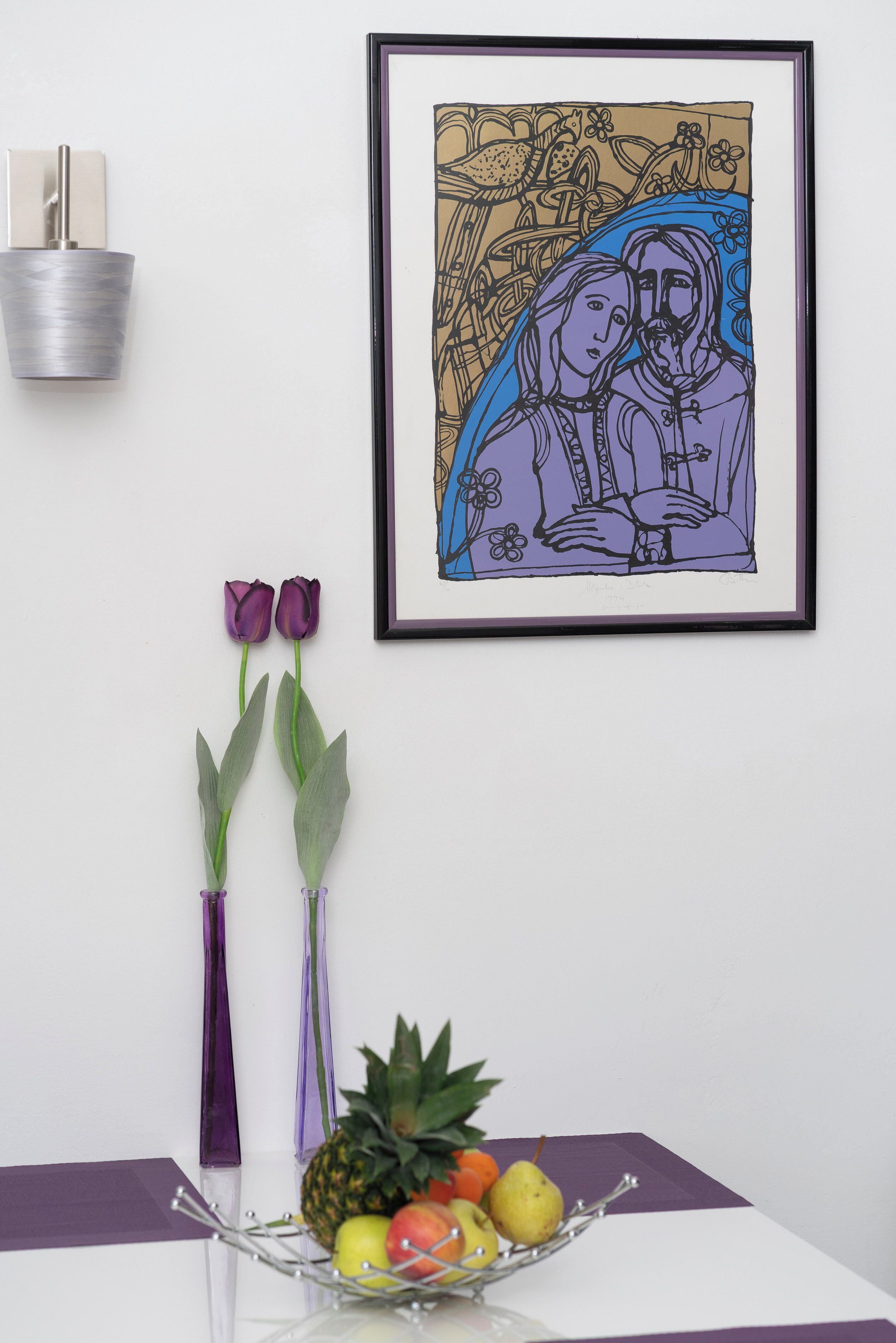 Ein gerahmtes Kunstwerk, zwei lilafarbene Vasen mit Blumen und eine Obstschale auf einem Tisch.