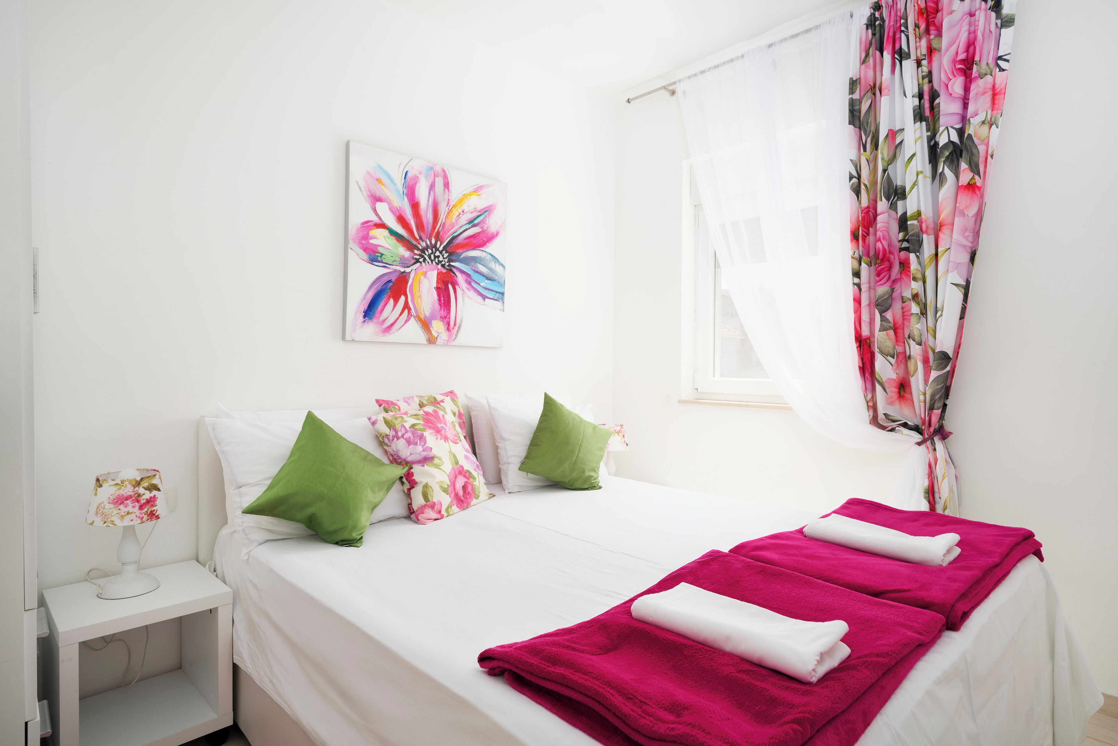 Una cama bien hecha con un juego de almohadas blanco y rosa floral, una almohada verde y una toalla rosa.