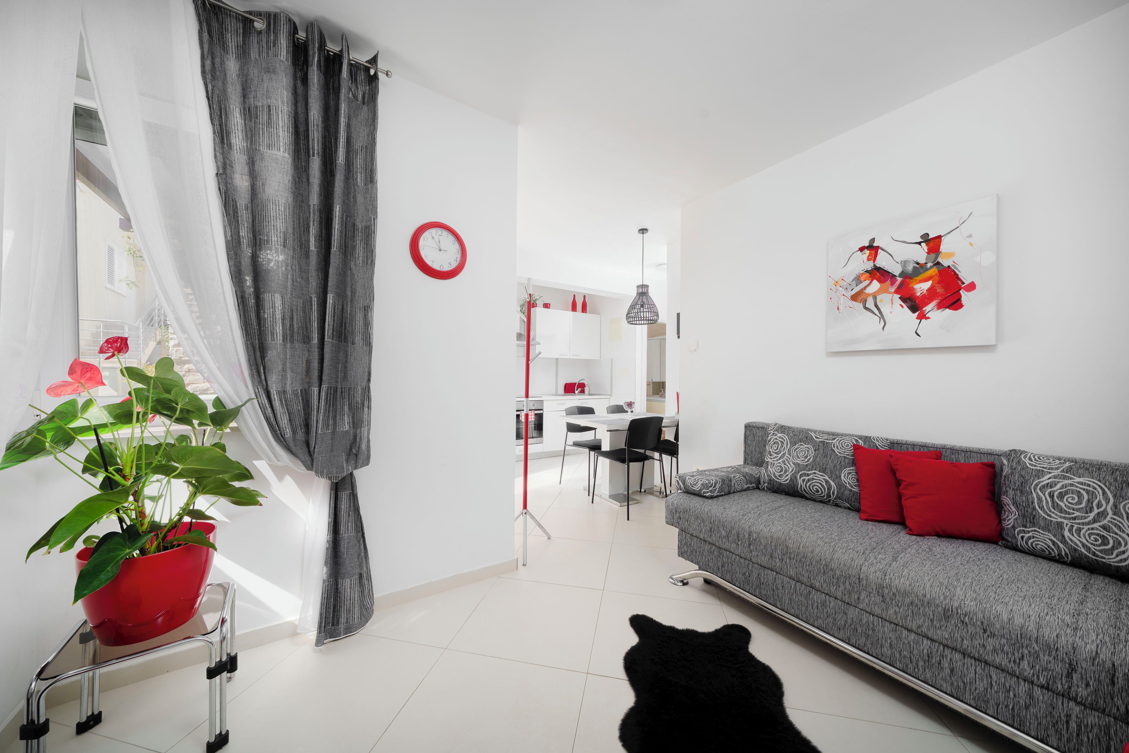 Ein modernes Wohnzimmer mit einem grauen Sofa, roten Kissen, einer schwarzen Katze, einem roten Topfpflanzen und einem Essbereich im Hintergrund.