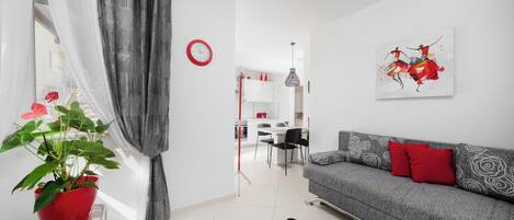 A modern living room with a grey sofa, red cushions, a black cat, a red potted plant, and a dining area in the background.