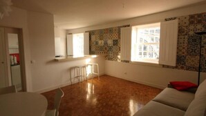 Een ruime woonkamer met een houten vloer, een witte bank, een eettafel met stoelen en een keuken met witte kastjes.