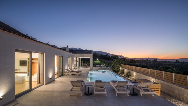A modern outdoor pool area with lounge chairs, a glass-enclosed patio, and a view of the mountains at dusk.