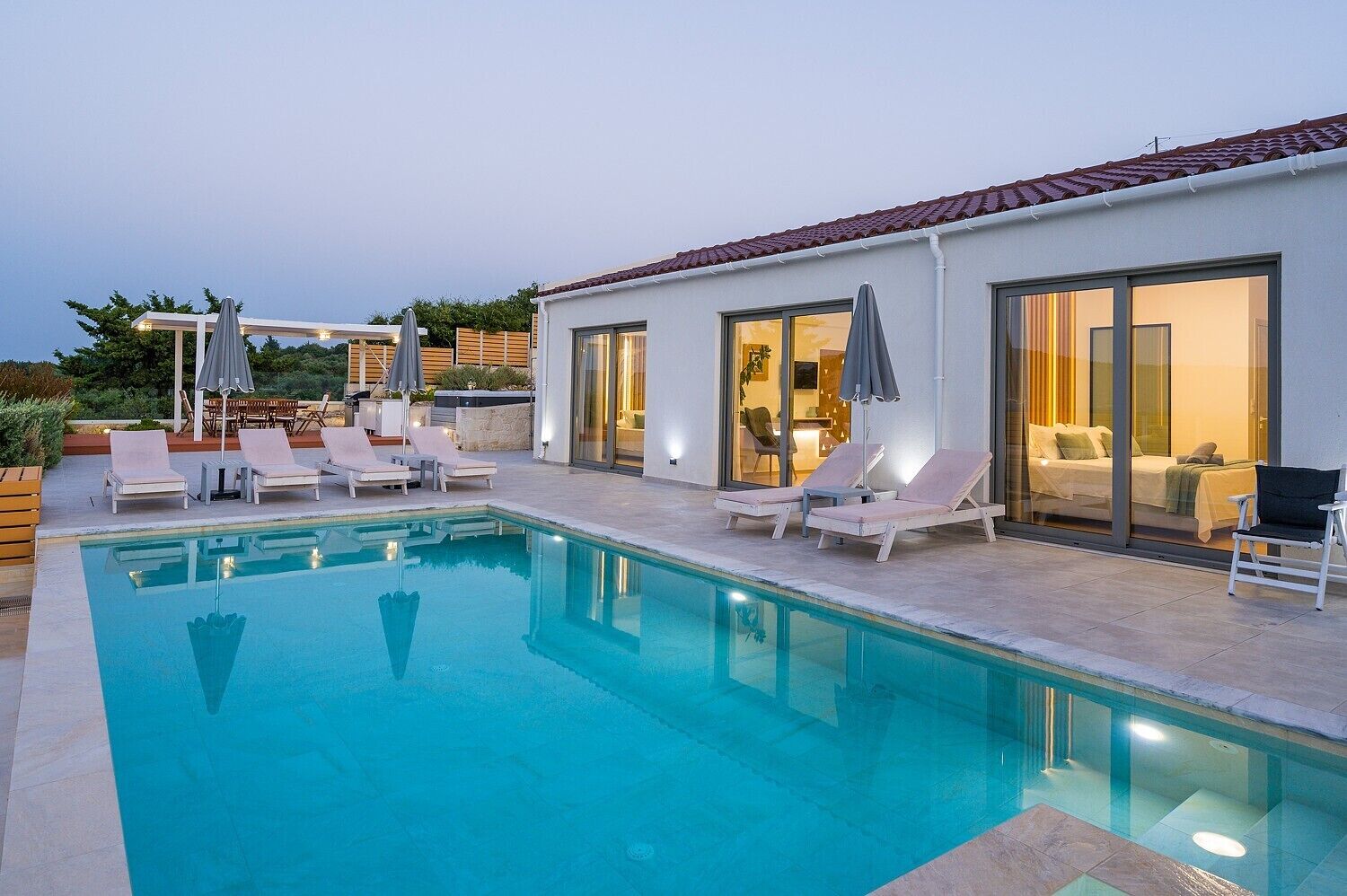 A modern outdoor pool area with lounge chairs and umbrellas, a well-lit interior, and a clear view of the pool.