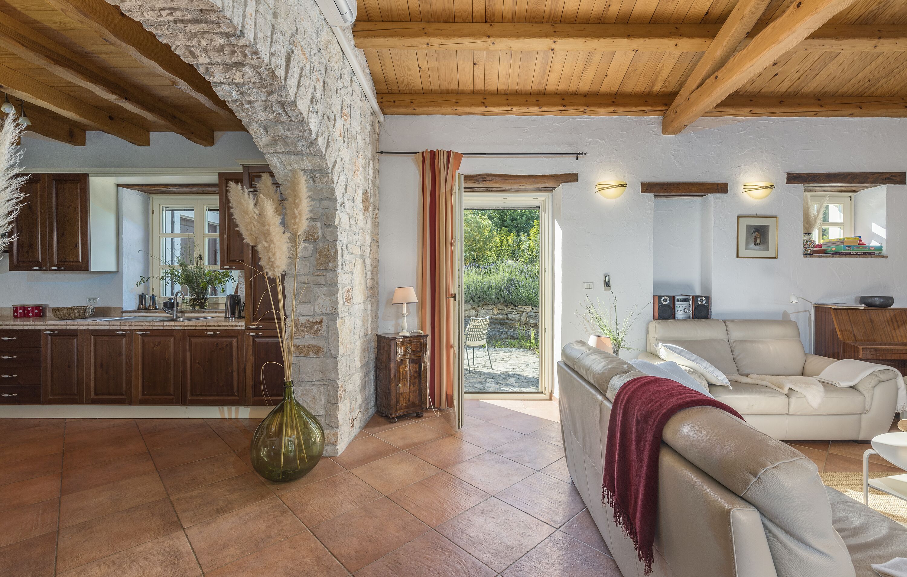 Ein geräumiger Wohnbereich mit einer Steinwand, Holzbalkendecke, einem beigefarbenen Sofa und einer Küche im Hintergrund.