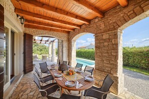 An outdoor dining area with stone arches, wooden beams, and a view of a swimming pool.