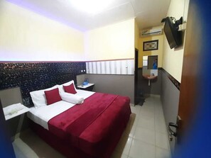 A hotel room with a bed, a nightstand, a TV mounted on the wall, and a bathroom visible through an open door. - Puri Made 2 Homestay (Banyuwangi)