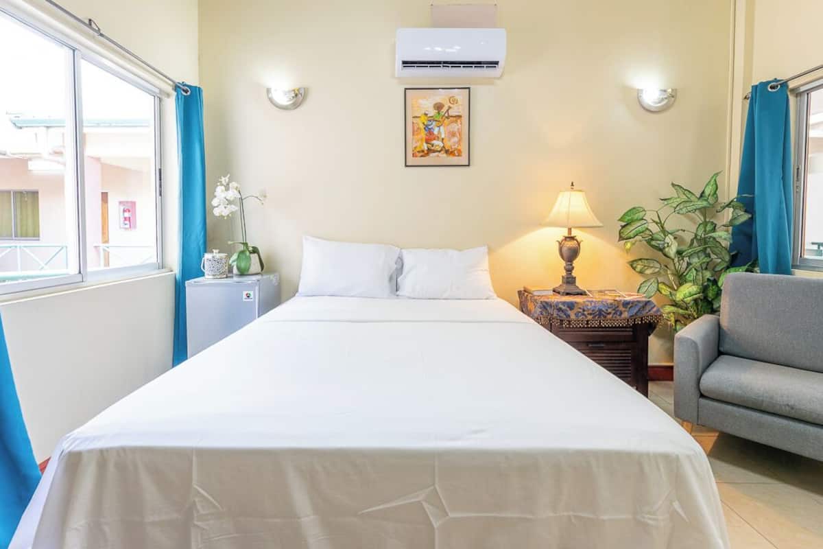 A hotel room with a bed, a nightstand with a lamp, a chair, a potted plant, and a window with curtains.