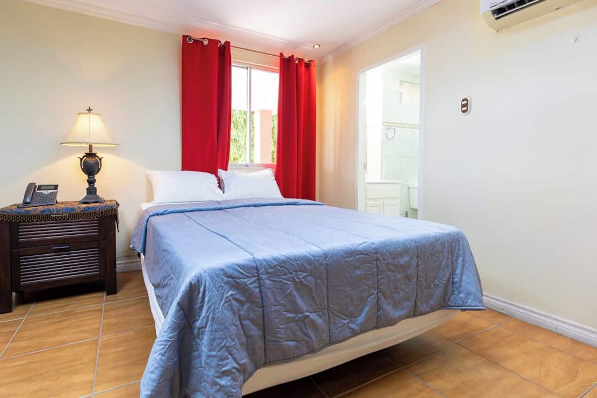 A bedroom with a bed, a nightstand, a lamp, and a window with red curtains.