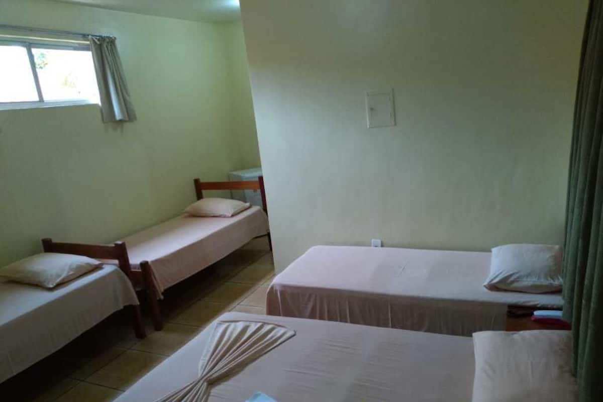 A room with three single beds, each with a wooden frame and white bedding. There is a window with curtains on the left side, and a power outlet on the wall.