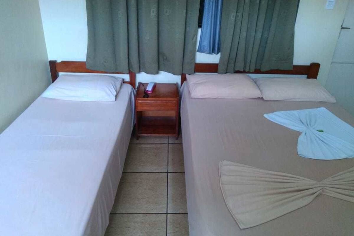 A room with two single beds, wooden headboards, a small bedside table with a lamp, and a window with curtains.