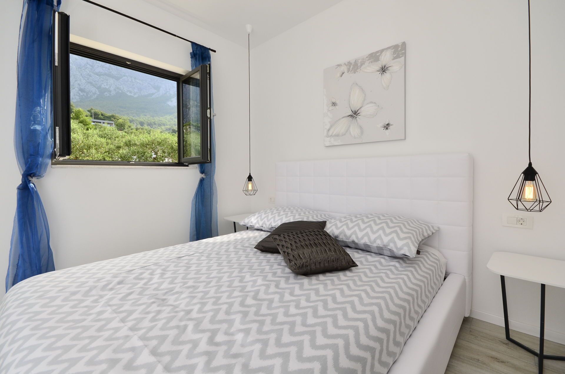 A bedroom with a bed, a window with curtains, a wall-mounted light fixture, and a painting.