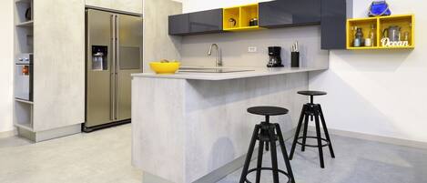 A modern kitchen with a central island, two bar stools, a built-in refrigerator, and a yellow shelf with kitchen items.