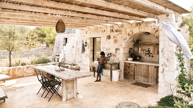 Un área de cocina al aire libre con pared de piedra, mesa de madera y un niño cerca de la entrada.