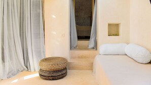 Una habitación pequeña y acogedora con una cama, un taburete tejido y una ventana con cortinas.