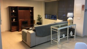 Una sala de estar con un sofá, una mesa de centro de vidrio, un mueble de madera, una planta en maceta y una ventana con persianas.