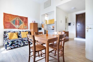 Un comedor con mesa y sillas de madera, un sofá con cojines de colores y una cocina al fondo.