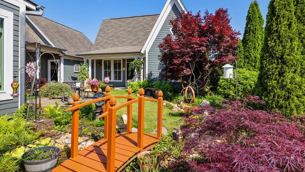 A residential property with a wooden bridge, a front porch, and a well-maintained garden with various plants and a tree with red leaves. - Clemson Lover's Dream Cottage (Clemson)