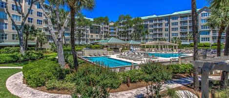 A resort with a central swimming pool surrounded by lounge chairs and palm trees.