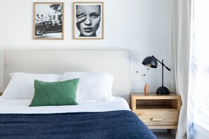 A neatly made bed with white and green pillows, a blue blanket, and a wooden nightstand with a lamp and a vase with flowers.