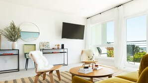 Ein modernes Wohnzimmer mit Sofa, Sessel und Couchtisch, einem flachbildfernseher an der Wand und einem großen Fenster mit Blick ins Freie.