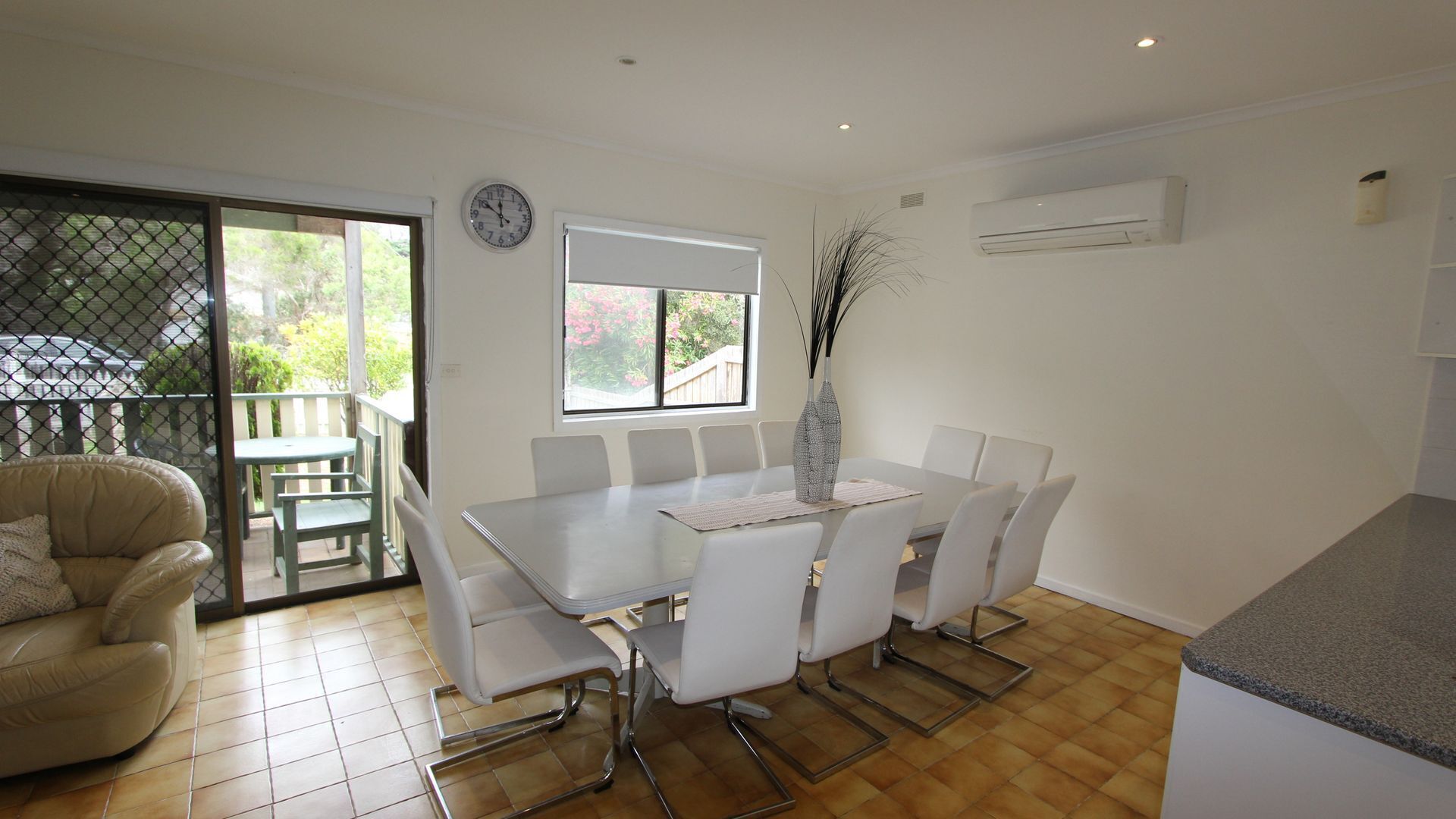 A dining area with a white table and chairs, a vase on the table, and a view of the outdoors through a window.