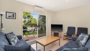 A living room with a sofa set, a coffee table, a TV, and a sliding glass door leading to an outdoor area with a table and chairs. - The Waves Sanctuary Apartment (COWES)