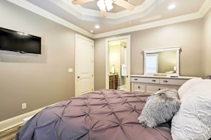 Un dormitorio con una cama grande, ventilador de techo, televisor montado en la pared y cómoda con espejo.