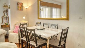 Un coin repas avec une table blanche et des chaises en bois, un grand miroir avec un cadre doré et une lampe posée sur une table d’appoint.