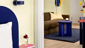 Ein Schlafzimmer mit einem Bett, einem Nachttisch mit Buch und Blumenvase, einem blauen Stuhl, einem kleinen Tisch mit Tasse und einem Sofa im angrenzenden Raum.