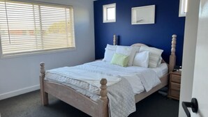 Une chambre à coucher avec un lit en bois, du linge de lit blanc, une fenêtre avec des stores et un mur bleu orné de deux tableaux encadrés.