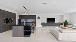 Et moderne køkken med indbyggede ovne, en central ø med barstole og et spiseområde med sofa og sofabord.