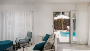 En moderne stue med sofa, stol, sofabord i glas og udsigt til et udendørs swimmingpool gennem en glasdør.