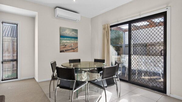 A modern dining area with a glass table and chairs, a painting of a beach scene, and a sliding glass door leading to an outdoor space.