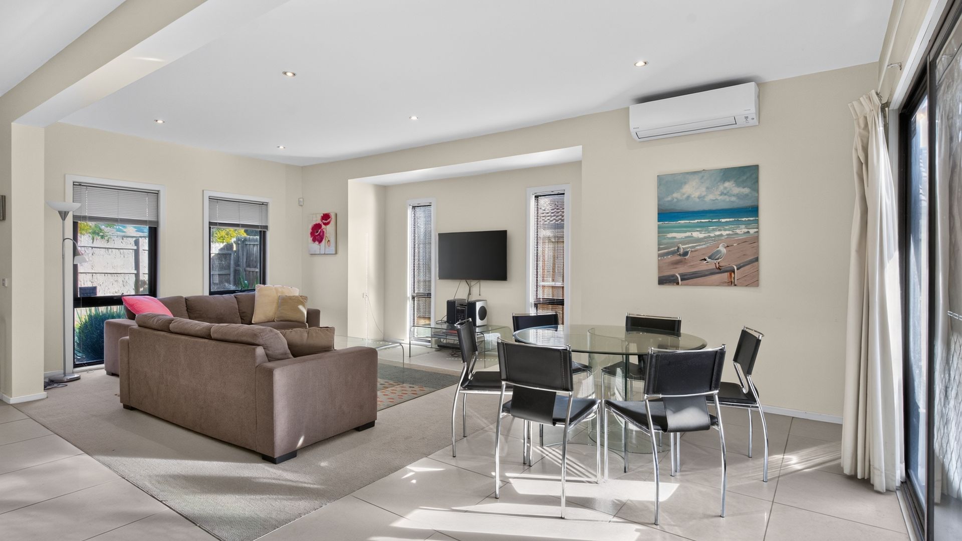 Een moderne woonkamer met een bank, een glazen eettafel met stoelen, een flatscreen televisie en een schilderij van een strand.