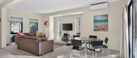 A modern living room with a sofa, a glass dining table with chairs, a flat-screen TV, and a painting of a beach scene.