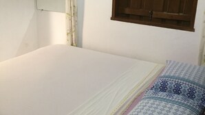 Une petite pièce simple, avec un lit simple, une fenêtre avec des volets en bois, un ventilateur fixé au mur et un miroir.