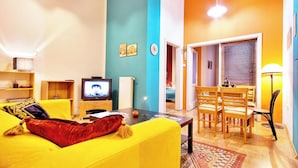 Ein Wohnzimmer mit gelbem Sofa, Fernseher und Essbereich mit Holztischen und Esstisch.