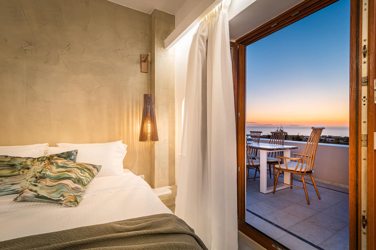 Una camera d'albergo con un letto, vista sul tramonto, un balcone con sedie e un tavolo.