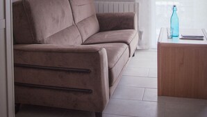 Una sala de estar con un sofá color beige, una pequeña mesa de centro de madera y una ventana con cortinas translúcidas.