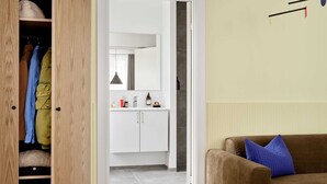 Une pièce avec une armoire en bois, un canapé marron avec un coussin bleu, et une vue sur une salle de bain équipée d’un miroir, d’un lavabo et d’articles de toilette.