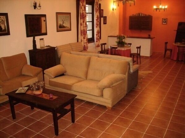 A living room with a beige sofa, a wooden coffee table, and a small side table. There is a dining area with a table and chairs in the background. - Rural house (full rental) El Empecinado for 23 people (Fuentelviejo)